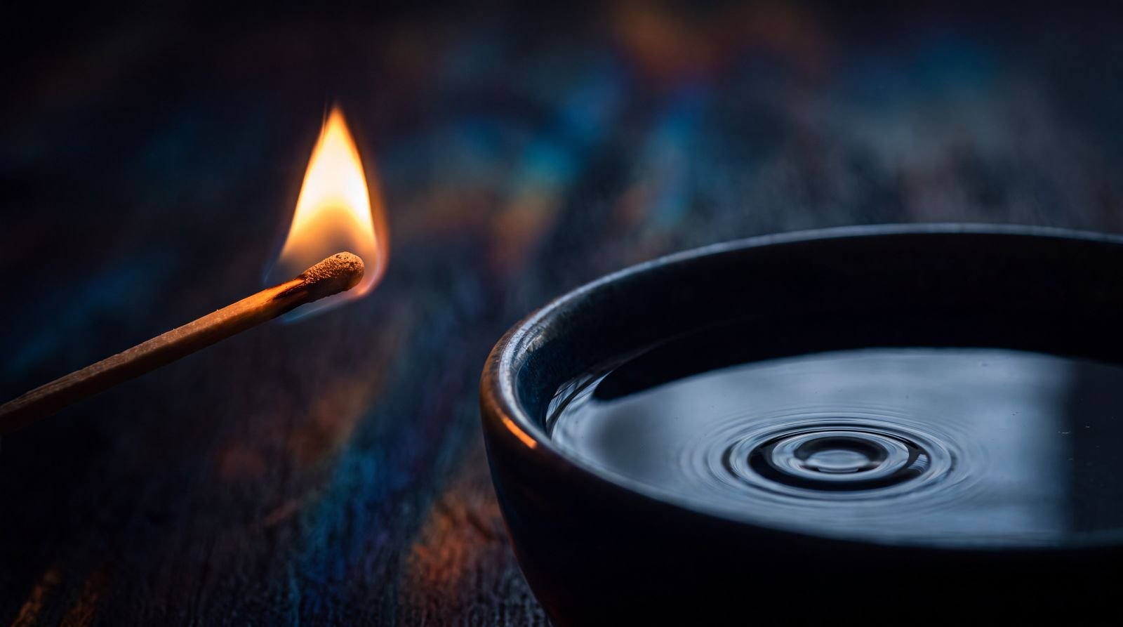 A lit match beside a bowl of still water with a single ripple, against a dark background